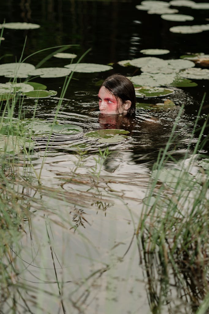 A mysterious figure with painted face emerges from lily pad-covered pond, evoking mythical themes.